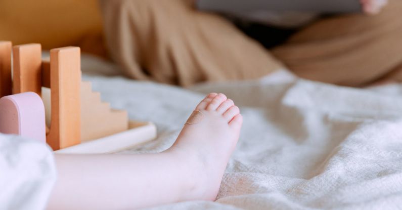 Remote Work Skills - Crop faceless toddler sitting on bed with colorful wooden building blocks while mother sitting with legs crossed doing remote work with laptop on knees on background