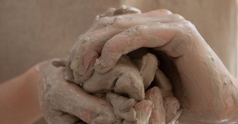 Industry Skills - Unrecognizable person wearing gray apron shaping and molding clay in dirty hands while making handicraft crockery in workshop during work