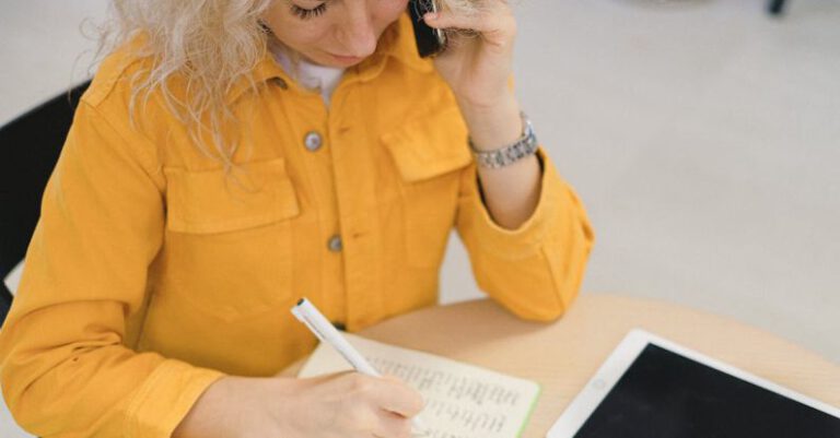 Job Fairs - From above of female freelancer taking notes in notebook while sitting at table and using smartphone for making call